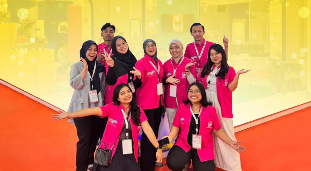 Group photo of smiling team members in pink shirts with lanyards posing together at an event or expo in a bright yellow backdrop