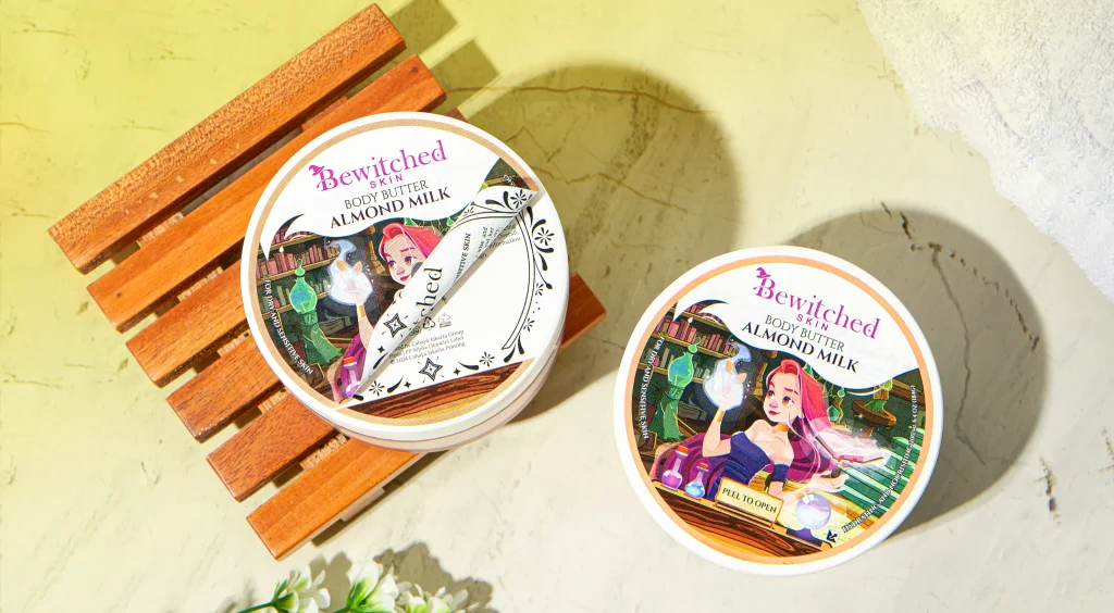 Two Bewitched Skin almond milk body butter tins resting on a wooden slatted bench atop a yellow surface.