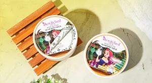 Two Bewitched Skin almond milk body butter tins resting on a wooden slatted bench atop a yellow surface.