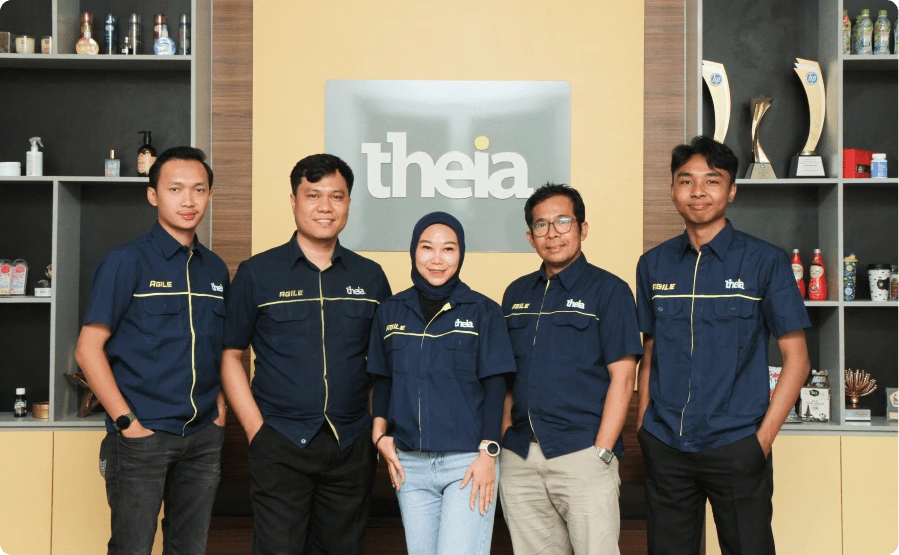 Five people in matching navy Theia uniforms pose for a group photo in front of a yellow wall with a Theia logo display.